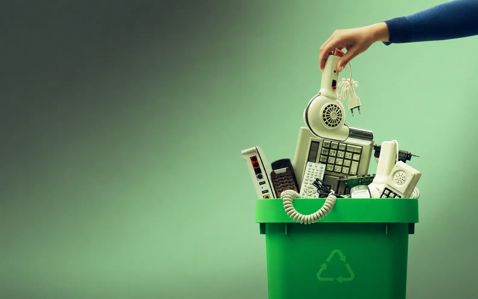 Mão colocando secador de cabelo em lixeira verde de reciclagem de lixo eletrônico com teclado, controles remotos, modem.